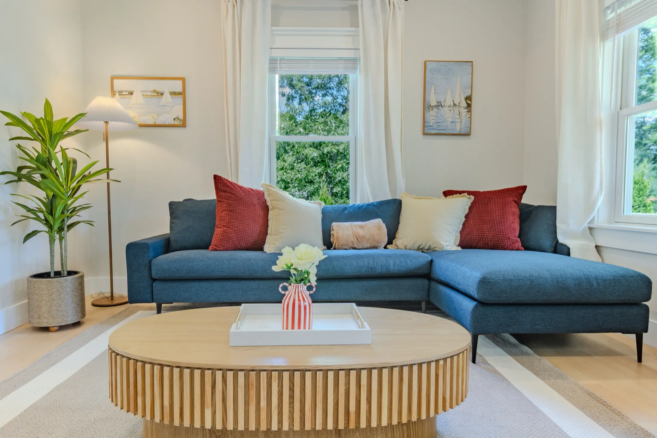 Living room with a deep blue sectional, rust and cream pillows, a round fluted oak coffee table, and tall windows onto trees.