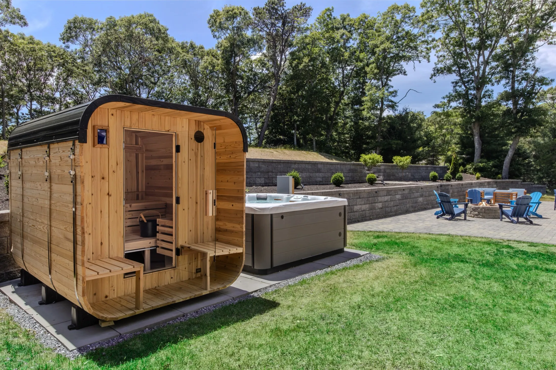 A barrel-shaped cedar sauna and a hot tub on a stone terrace, with a fire pit and blue Adirondack chairs beyond.